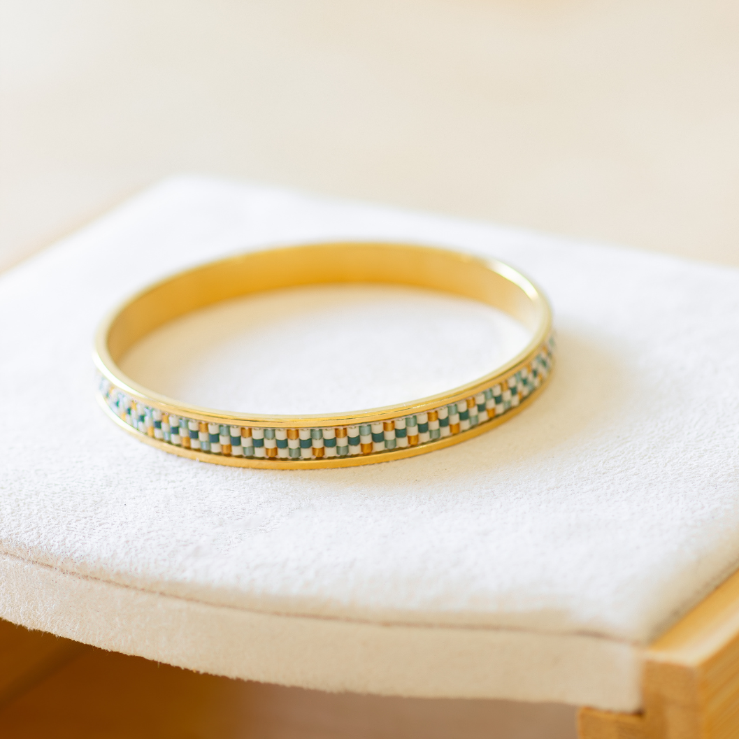 Hand-beaded Tatreez bangle in marigold, sea glass green, evergreen and dark cream on white background. Three-row Miyuki glass bead pattern on 304 stainless steel.